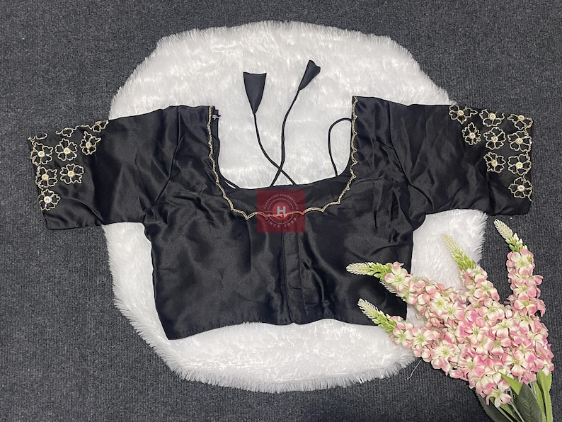 May include: A black blouse with short sleeves featuring gold floral embroidery on the cuffs and neckline. The garment has a square neckline and thin black tie strings at the back. The fabric has a smooth, satin-like finish with a subtle sheen.