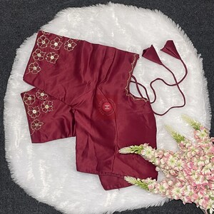 May include: A deep maroon blouse featuring short sleeves with gold floral embroidery and sequin accents. The garment includes thin matching fabric ties with cone-shaped ends at the side and a subtle scalloped edge detail along the sleeve hem.