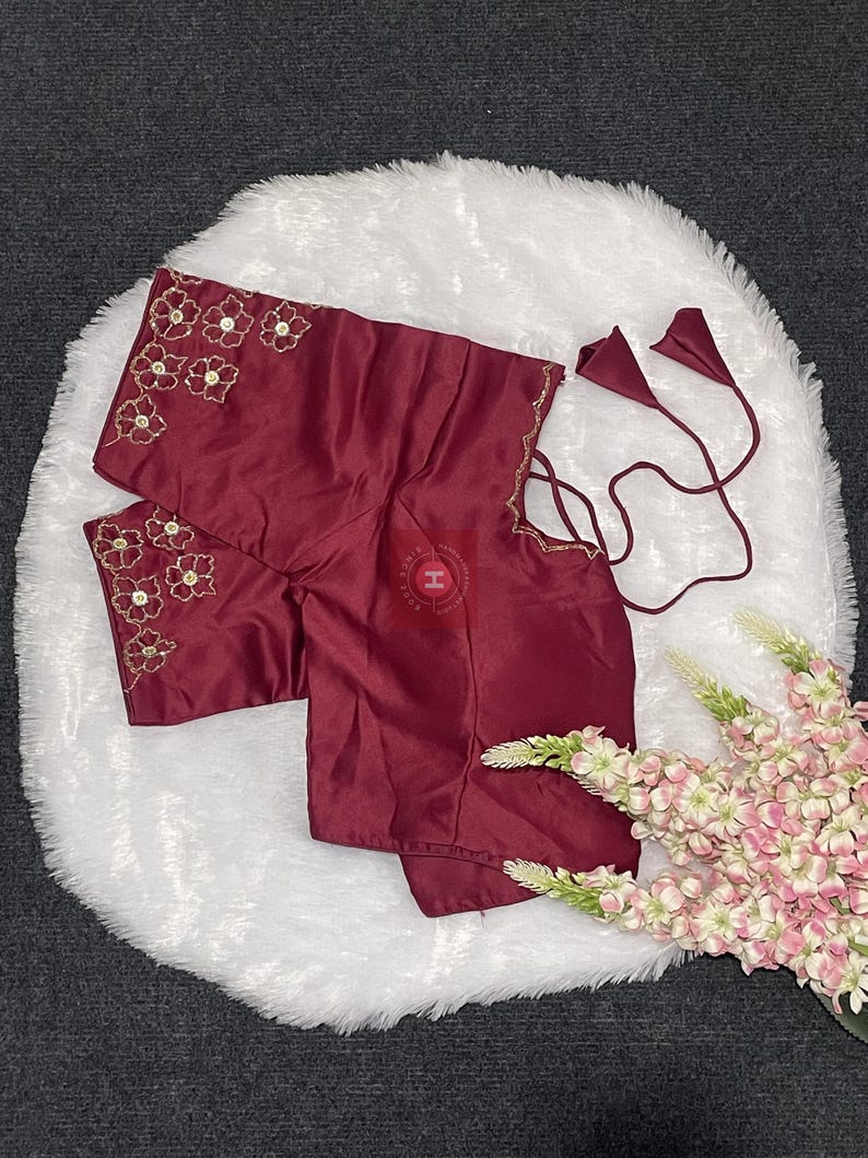 May include: A deep maroon blouse featuring short sleeves with gold floral embroidery and sequin accents. The garment includes thin matching fabric ties with cone-shaped ends at the side and a subtle scalloped edge detail along the sleeve hem.