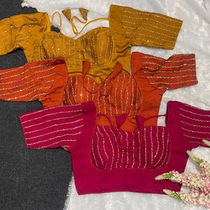 Può includere: Tre camicette a maniche corte in giallo senape, arancione bruciato e magenta. Ogni camicetta presenta uno scollo a cuore, file verticali di ricami metallici argentati sul corpetto e sulle maniche, e sottili lacci in tessuto abbinati sul retro.