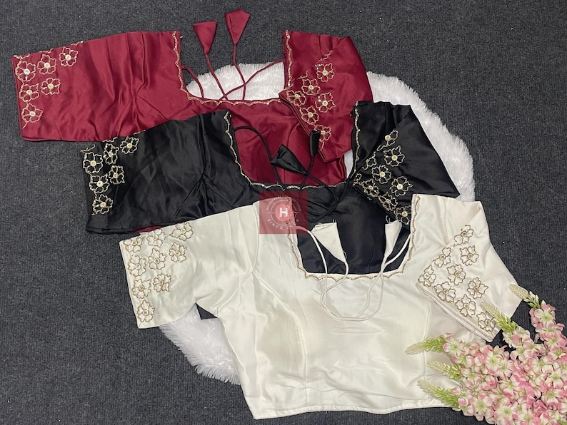 May include: Three short-sleeved blouses in maroon, black, and white, each featuring a square neckline and floral embroidery on the sleeves. The garments include thin tie-up strings at the back and a small red square logo in the center.