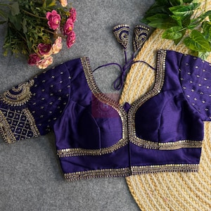 May include: A deep purple blouse with gold embroidery and embellishments. The blouse features a V-neckline and short sleeves, with intricate detailing around the neckline and sleeves. The design includes small, gold-colored sequins and a decorative pattern.