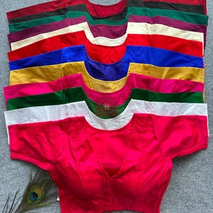 May include: A stack of short-sleeved blouses in various solid colours including hot pink, white, dark green, yellow, blue, red, cream, purple, and black. Each blouse features a V-neckline and a structured bodice design, arranged in a vertical column.