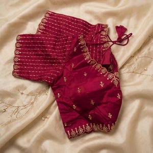 May include: A maroon blouse with intricate gold embroidery. The blouse features short sleeves with horizontal gold lines and a scalloped edge. The bodice is adorned with delicate floral patterns and a drawstring closure. The garment is displayed on a cream-colored fabric.