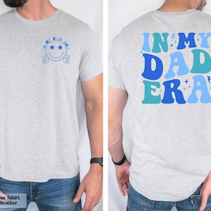 May include: Light gray t-shirt with blue text and graphics. The front features a smiley face with the words "IN MY DAD ERA". The back of the shirt has the words "IN MY DAD ERA" in a retro font. Athletic heather fabric.