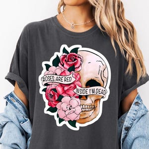 May include: Dark gray t-shirt featuring a skull graphic with pink roses and the text "Roses are red, inside I'm dead." The design is a mix of floral and gothic elements, suitable for casual wear.