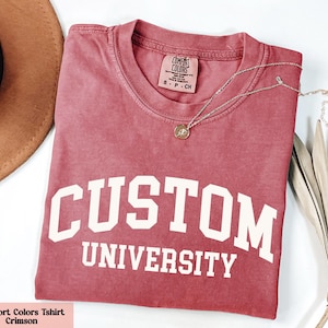 May include: Crimson Comfort Colors t-shirt with the words "CUSTOM UNIVERSITY" in white block letters. A gold necklace with a pendant rests on the shirt. A brown hat is visible in the upper left corner.