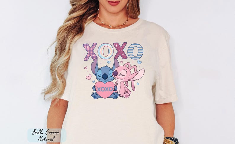 A woman wearing a white t-shirt with a graphic design featuring two cartoon characters, a blue character and a pink character, holding a heart and surrounded by the text "XOXO".