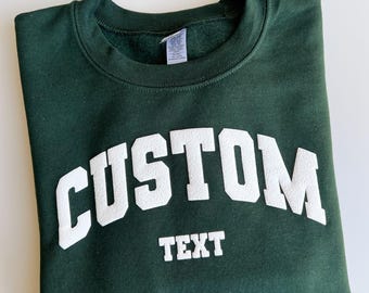 May include: A forest green Gildan sweatshirt with the word "CUSTOM" arched across the chest in white block letters, with "TEXT" below. The sweatshirt is folded neatly on a white surface.