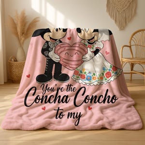 A pink blanket with a cartoon image of Mickey Mouse and Minnie Mouse holding a heart, along with the text "You're the Concha Concho to my" printed on it.