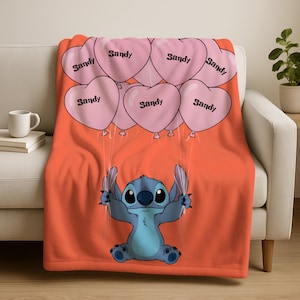An orange blanket with a cartoon character, specifically Stitch, sitting underneath pink heart-shaped balloons with the word "Sandy" written on them.