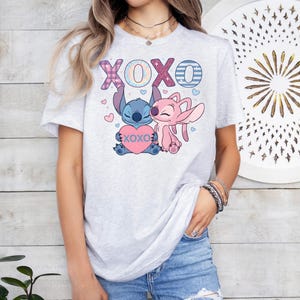A woman wearing a white t-shirt with a graphic design featuring two cartoon characters, one pink and one blue, along with the text "XOXO" and a heart.