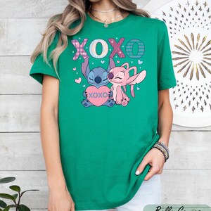 A woman wearing a green t-shirt with a graphic design featuring two cartoon characters, one pink and one blue, along with the text "XOXO" and hearts.
