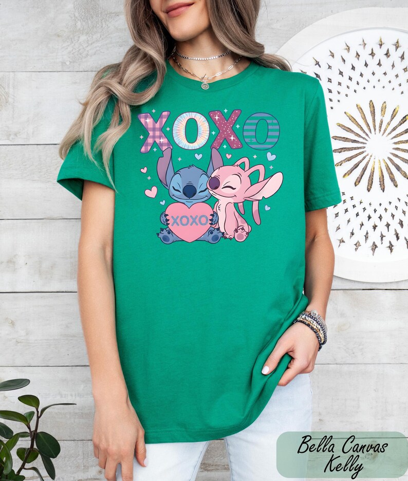 A woman wearing a green t-shirt with a graphic design featuring two cartoon characters, one pink and one blue, along with the text "XOXO" and hearts.