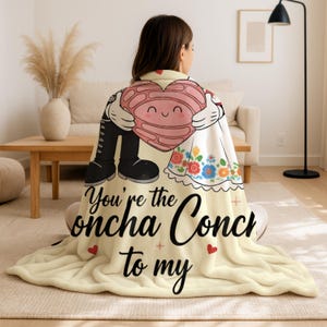 A woman sitting on the floor with a blanket featuring a cartoon character holding a heart and the text "You're the Onchka Conch to my".
