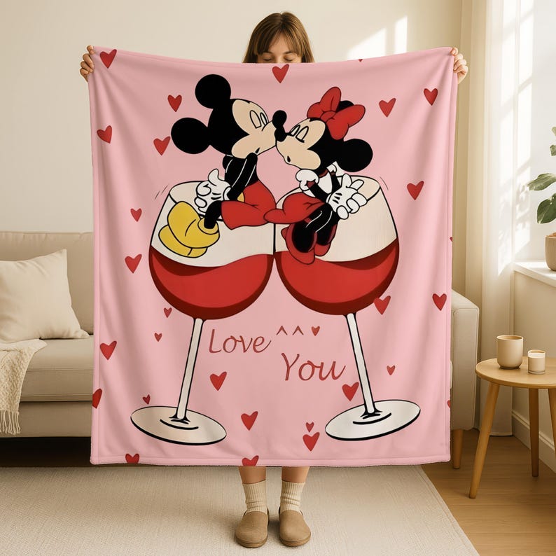 A woman is holding a pink blanket with a cartoon image of Minnie Mouse and Mickey Mouse kissing inside wine glasses, surrounded by hearts.