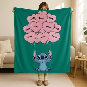 A person is holding up a large green blanket with a cartoon character, specifically Stitch, on it. The blanket has pink heart-shaped balloons with the word "Sandy" written on them.