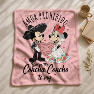 a pink blanket with a cartoon illustration of Mickey Mouse and Minnie Mouse, along with a Spanish phrase "Amor Prohibido" and a heart symbol.