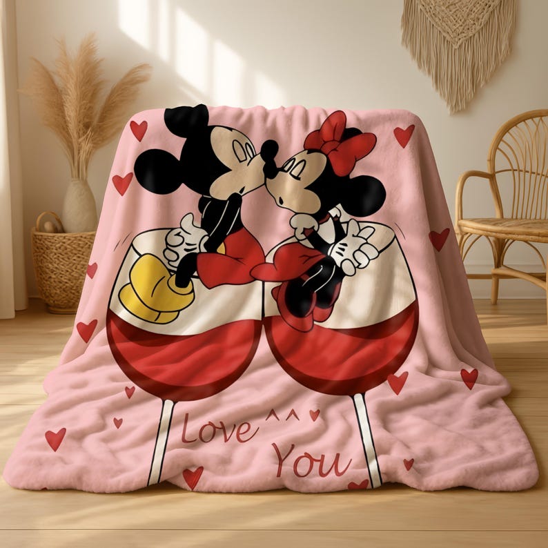 A pink blanket with a cartoon image of Minnie Mouse and Mickey Mouse sitting together and kissing, surrounded by hearts.