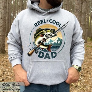 A man wearing a grey hoodie with a graphic design of a fish and the text "REEL. COOL. DAD" printed on it, standing in a forested area.