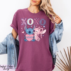 A woman wearing a purple t-shirt with a graphic of two cartoon characters, Stitch and Lilo, and the text "XOXO" in a pink color. She is also wearing a denim jacket and has her hair in a ponytail.