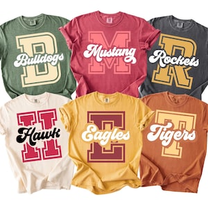 May include: Six different colored t-shirts with team names and logos. The shirts are in shades of green, red, gray, cream, yellow, and orange. Each shirt features a large letter with a team name in a retro font.