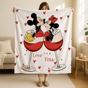 A woman is holding up a blanket with a cartoon image of Minnie Mouse and Mickey Mouse kissing inside wine glasses, surrounded by red hearts.