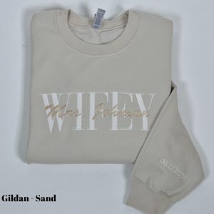 May include: Sand-colored sweatshirt with the word "WIFEY" embroidered in white, and "Mrs. Johnson" in a cursive font. The sleeve features the date "06.12.2024" embroidered in white. The sweatshirt is folded.