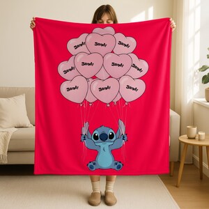 A woman is holding up a red blanket with a cartoon character, specifically Stitch, on it. The blanket has a design of pink heart-shaped balloons with the word "Sandy" written on them.