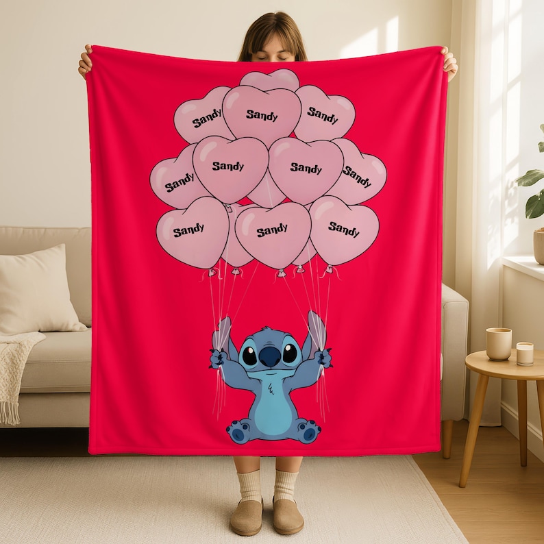 A woman is holding up a red blanket with a cartoon character, specifically Stitch, on it. The blanket has a design of pink heart-shaped balloons with the word "Sandy" written on them.