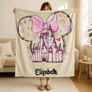 A woman is holding up a beige blanket with a drawing of a castle and a large pink bow on it.