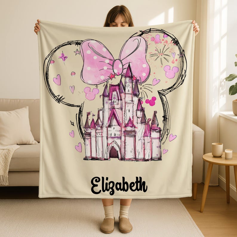 A woman is holding up a beige blanket with a drawing of a castle and a large pink bow on it.