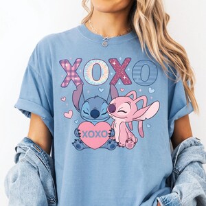 A woman wearing a blue t-shirt with a graphic of two cartoon characters, one pink and one blue, and the text "XOXO" in pink and blue. She is also wearing a denim jacket and denim shorts.