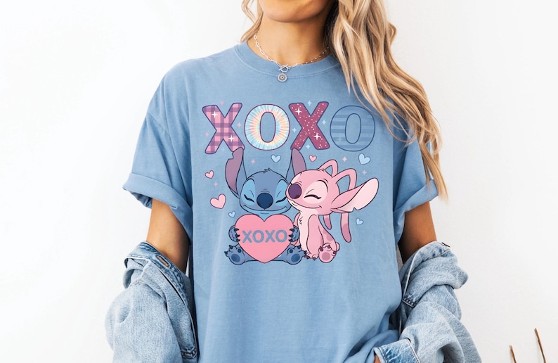 A woman wearing a blue t-shirt with a graphic of two cartoon characters, one pink and one blue, and the text "XOXO" in pink and blue. She is also wearing a denim jacket and denim shorts.