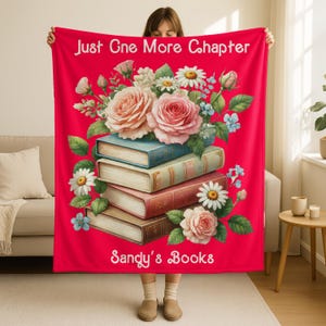 May include: A red blanket with the text "Just One More Chapter" and "Sandy's Books." The blanket features a floral design with roses, daisies, and books. The books are stacked and have blue, red, and brown covers.