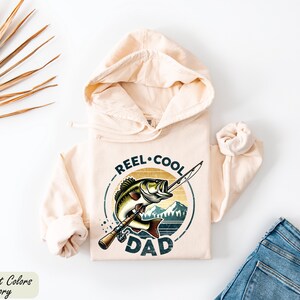 A beige hoodie with a graphic design of a fish and the text "REEL. COOL. DAD" printed on it, along with a pair of jeans and some dried leaves.