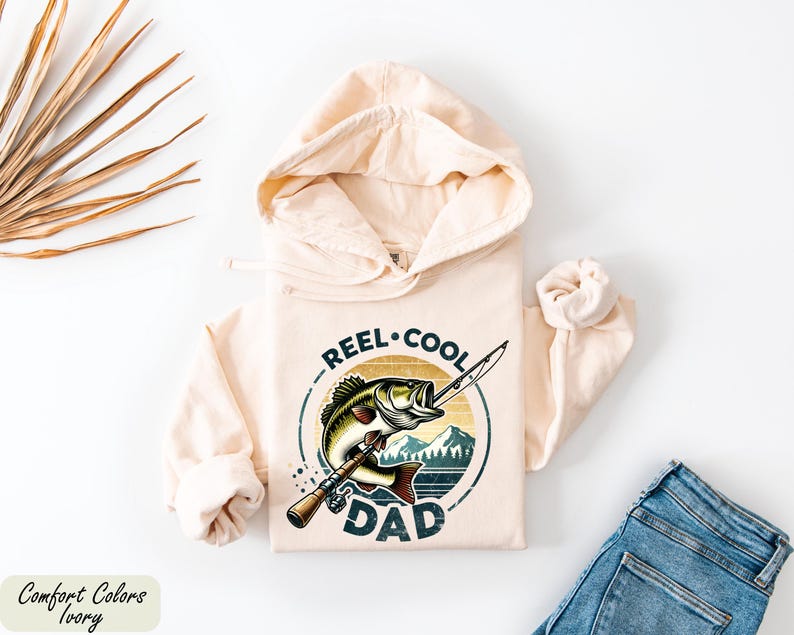 A beige hoodie with a graphic design of a fish and the text "REEL. COOL. DAD" printed on it, along with a pair of jeans and some dried leaves.