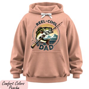A peach-colored hoodie with a graphic design featuring a fish and the text "REEL-COOL DAD" printed on it.