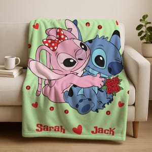 A green blanket with a cartoon image of two characters, a pink bunny and a blue cat, sitting together and holding a bouquet of flowers. The blanket has the names 'Sarah' and 'Jack' printed on it.