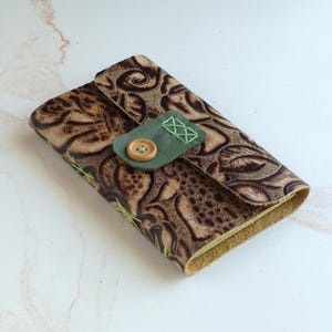 May include: A rectangular leather wallet featuring a dark brown embossed floral and textured pattern. The wallet is secured with a sage green leather flap closure, accented by a single light brown wooden button and decorative light green cross-stitching.