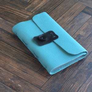 May include: A rectangular journal with a textured turquoise cover. The journal features a flap closure secured by a dark brown leather tab and a single black button. The spine and edges show visible stitching details along the turquoise material.