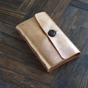 May include: A rectangular wallet with a metallic gold textured finish. The wallet features a single flap closure secured by a large, round black button with a small red thread detail visible in the center. The edges show red stitching.