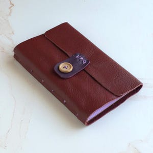 May include: A rectangular journal bound in dark maroon leather with a textured surface. The cover features a flap secured by a dark purple leather tab and a single light brown wooden button. The spine is stitched with light purple thread.