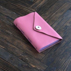 May include: A pink leather wallet with a triangular flap closure secured by a single white button. The wallet features visible purple cross-stitch detailing along the side and flap, with a smooth texture and a folded construction style.