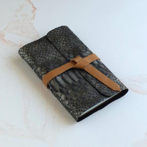 May include: A rectangular journal with a dark gray and black snakeskin pattern cover. The journal is secured by a tan leather strap that wraps around the center and ties in a knot on the front cover, keeping the pages closed and protected.
