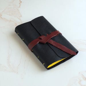 May include: A rectangular journal with a textured black cover, secured by a dark maroon leather wrap tie. The spine features visible yellow stitching along the edge, and the pages inside are bright yellow, visible at the side of the closed book.