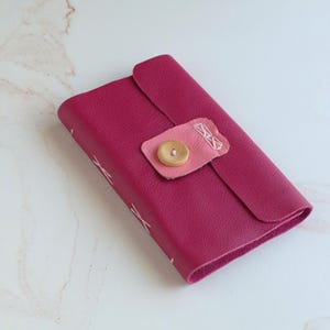 May include: A rectangular fuchsia wallet with a textured surface, featuring a light pink leather tab closure secured by a single round wooden button. The edges are finished with white cross-stitch detailing along the side and the closure tab.