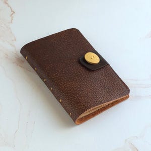 May include: A dark brown textured leather journal with a rectangular flap closure secured by a single round yellow button. The spine features visible yellow stitching along the edge, and the interior pages are visible at the bottom opening.