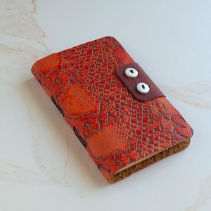May include: A rectangular notebook cover featuring a vibrant red and orange snakeskin pattern. A dark brown leather tab secures the cover, accented with two white circular buttons that have black centers, resembling a pair of stylized eyes.