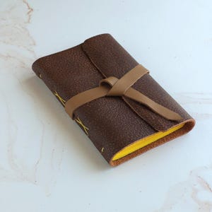 May include: A small, rectangular journal with a dark brown, textured leather cover. The journal is secured by a light tan leather wrap-around tie. The pages are bright yellow, and the binding features visible yellow cross-stitch detailing.